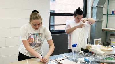 two students working on sculpture projects at a large work table.