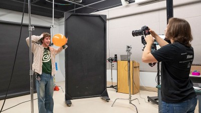 A student holding a balloon in a photo studio has his picture taken.
