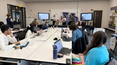 a group of students presents at the front of the room while other students watch from a long table.