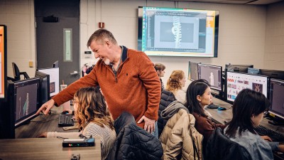 Faculty Craig Foster points at a computer in a lab where students are working.