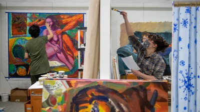 two students painting in a studio space, with one student's work depicting a nude woman in pinks and purples.