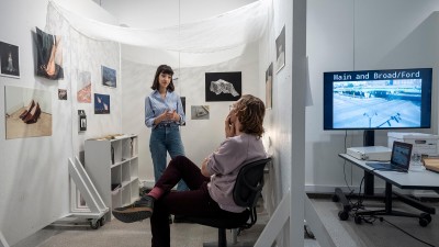 student and professor talk in a small gallery space with sheer fabric hanging from the ceiling.