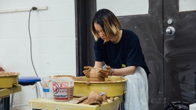 a student spins a tall clay vessel on a wheel.