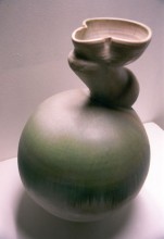 A sage green-glazed ceramic jar that's rounded and abstract in shape. The neck of the jar is narrowed and angled upward diagonally to where it opens at the top.
