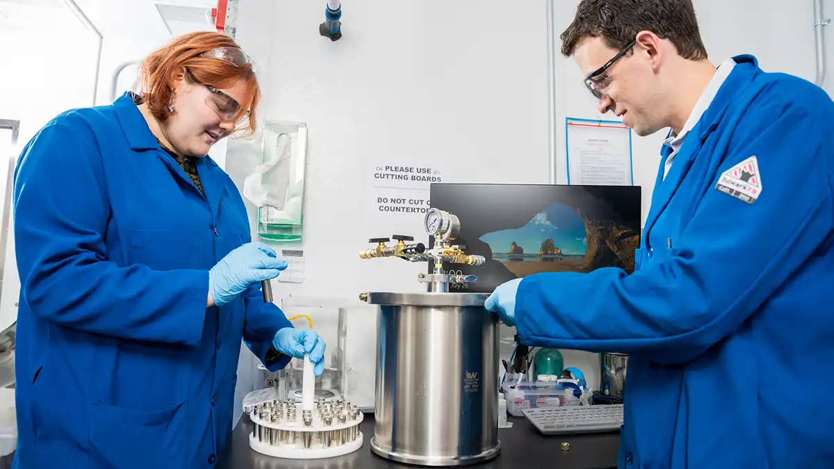 Two researchers in blue lab coats handle battery materials in a laboratory environment.