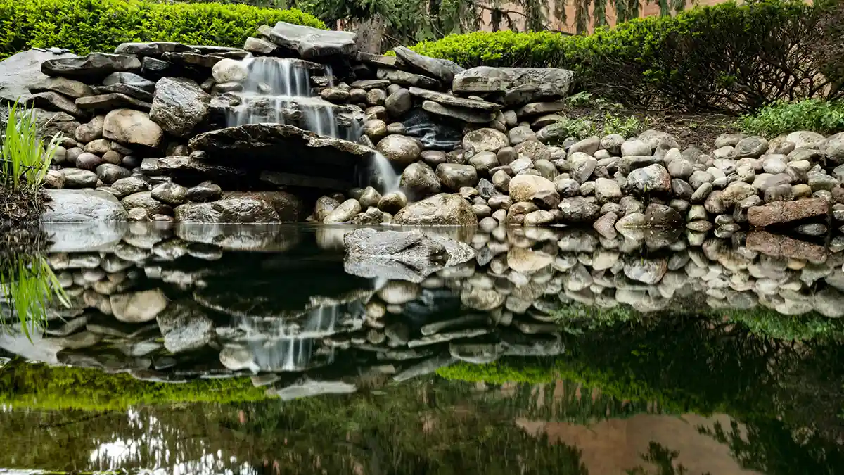 A small waterfall flows over rocks into a calm pond surrounded by greenery.