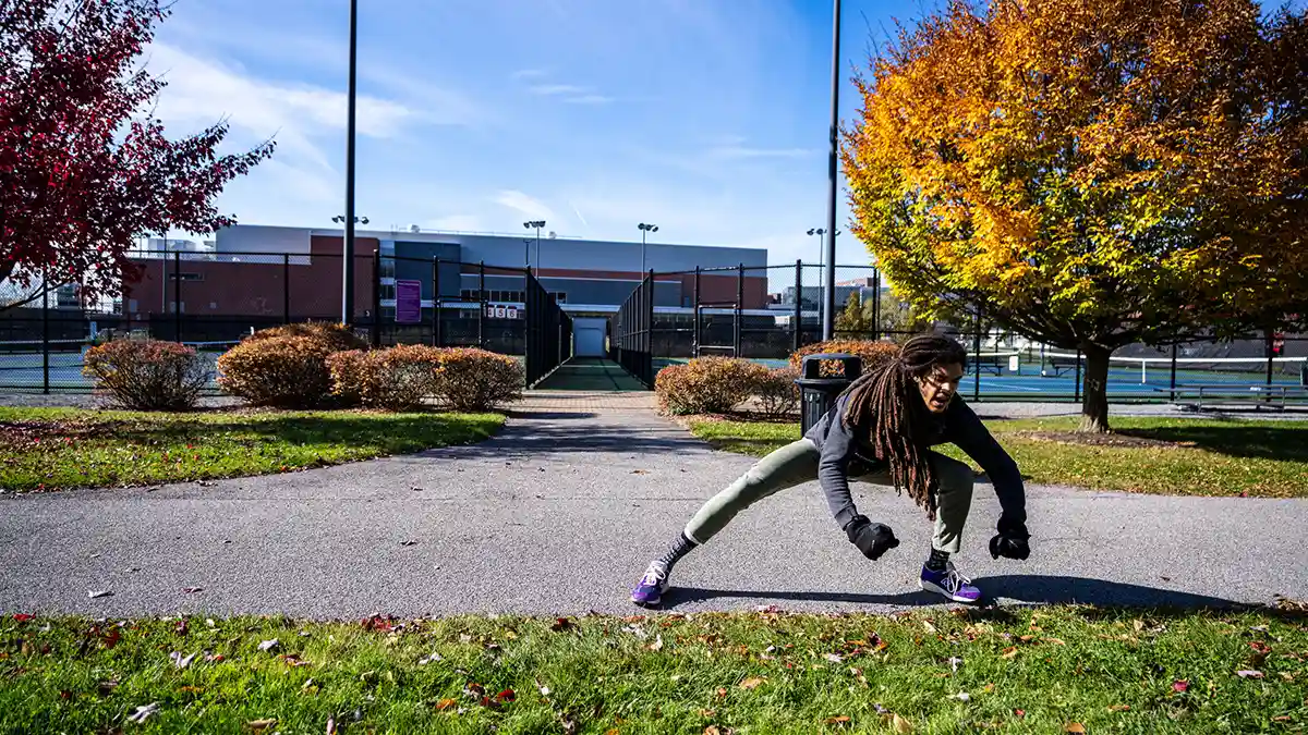 A person stretches outdoors near tennis courts with colorful autumn trees in the background.