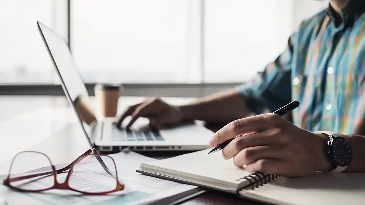 A person works on a laptop while writing notes beside documents and glasses.