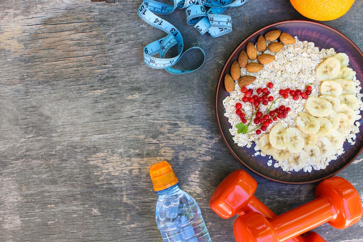 A bowl of oatmeal with fruit, dumbbells, a water bottle and tape measure on a table.