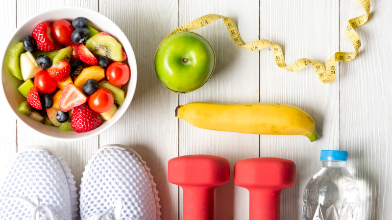 A bowl of fruit, running shoes, an apple, dumbbells, and water bottle on a table