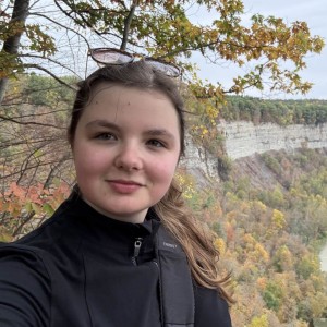 Person taking a selfie at a scenic overlook with a winding river and forested cliffs.
