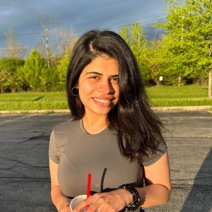 A girl smiling outdoors, holding a drink, with greenery in the background.