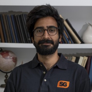 Professional headshot of Shahzeb wearing a black student government t-shirt with a bookshelf background