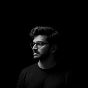 Portrait of a Melson with glasses looking to the side, dramatically lit against a black background, highlighting only part of his face and upper body.