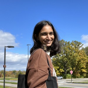 A young woman with a joyful smile, standing outdoors on a sunny day with autumn trees in the background. She wears a brown jacket and carries a black tote bag, embodying a casual, relaxed look.