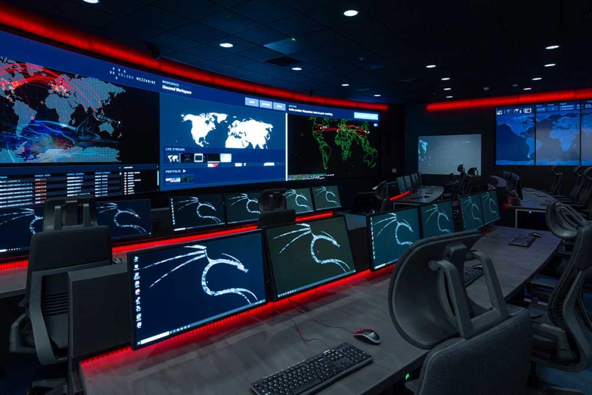 A modern cybersecurity operations center with rows of computer workstations facing large wall-mounted displays. Multiple screens show world maps, network monitoring dashboards, and data visualizations, while red accent lighting illuminates the dark.