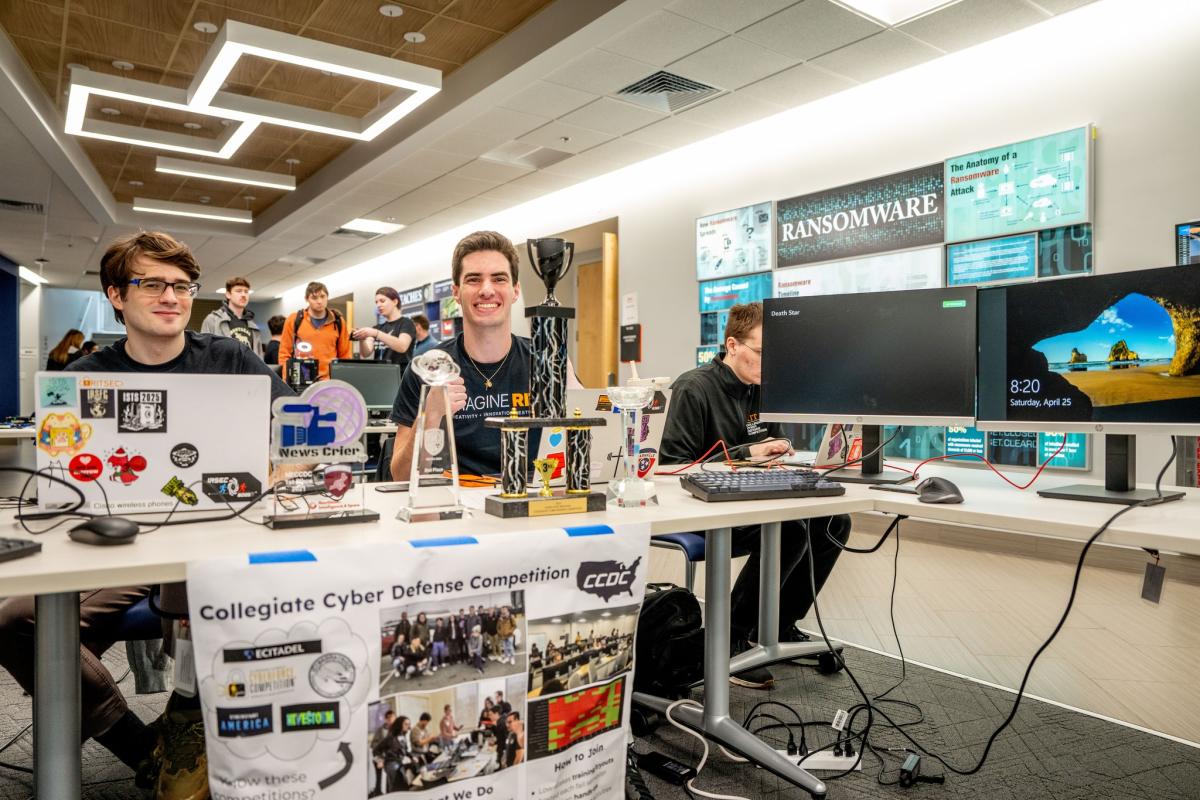 Students staff a RITSEC cybersecurity exhibit with laptops, monitors, competition trophies, and information displays during a technology showcase event.