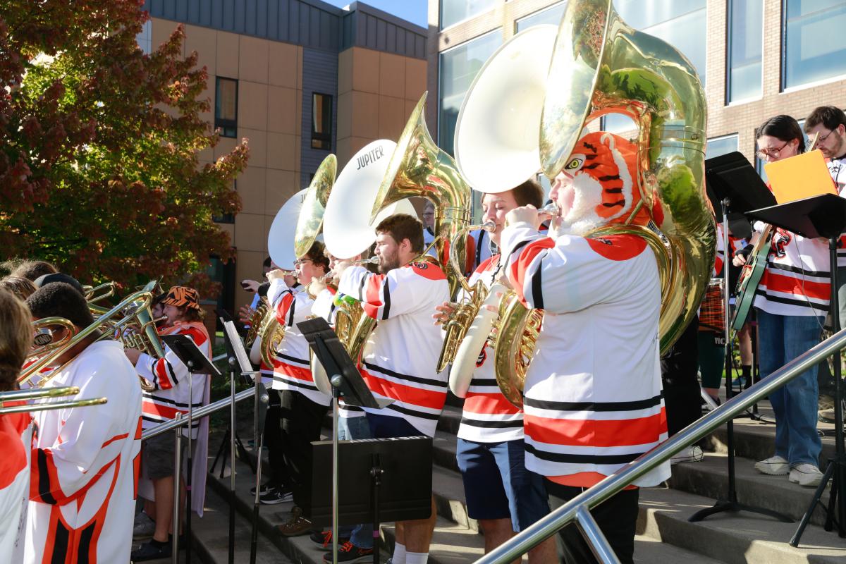 RIT Pep Band members perform outdoors, playing brass instruments while wearing hockey jerseys.