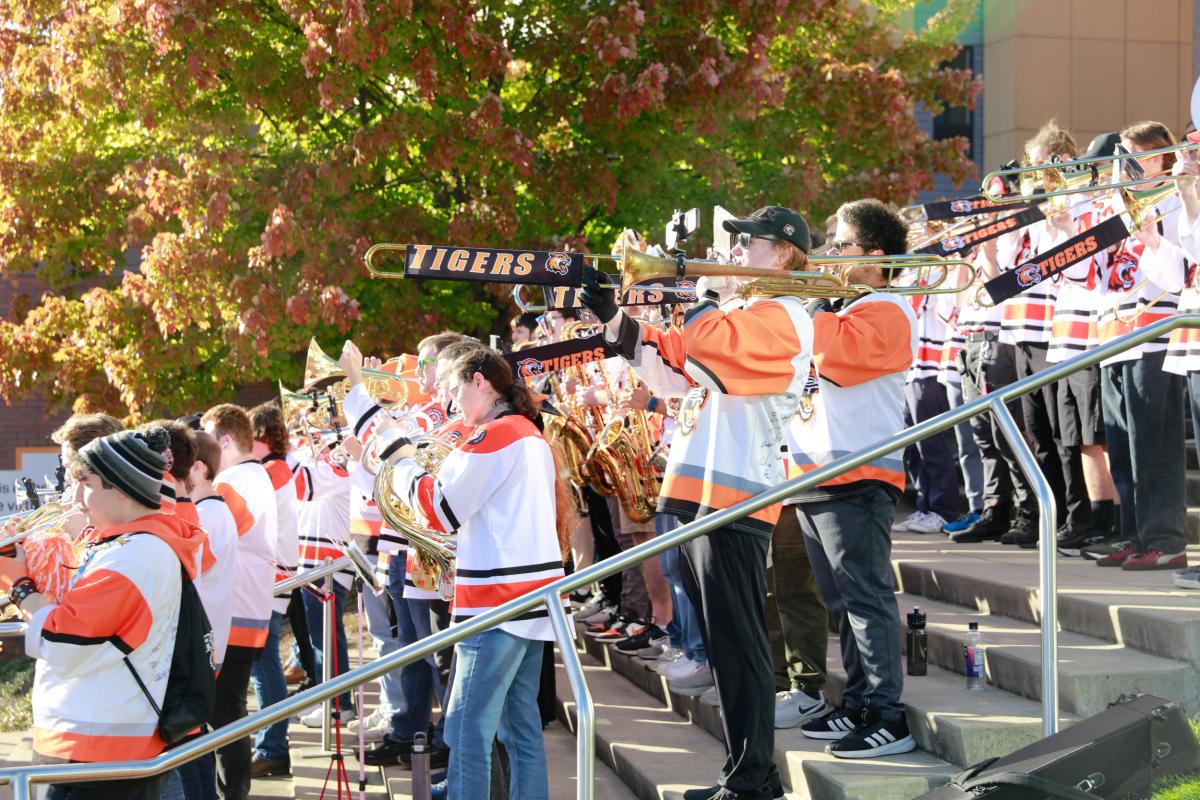 RIT Pep Band members perform outdoors on campus steps, playing brass instruments while wearing team jerseys.
