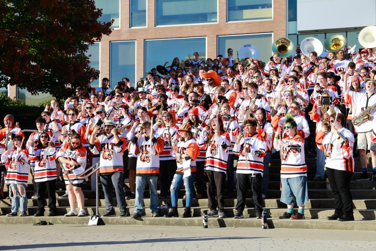 RIT Pep Band members gather on outdoor campus steps, performing in matching hockey jerseys.