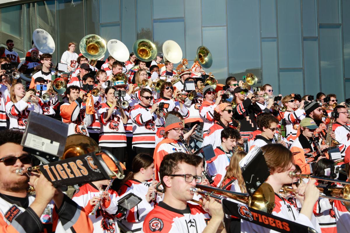 RIT Pep Band members perform outdoors, playing brass and woodwind instruments while wearing team jerseys.