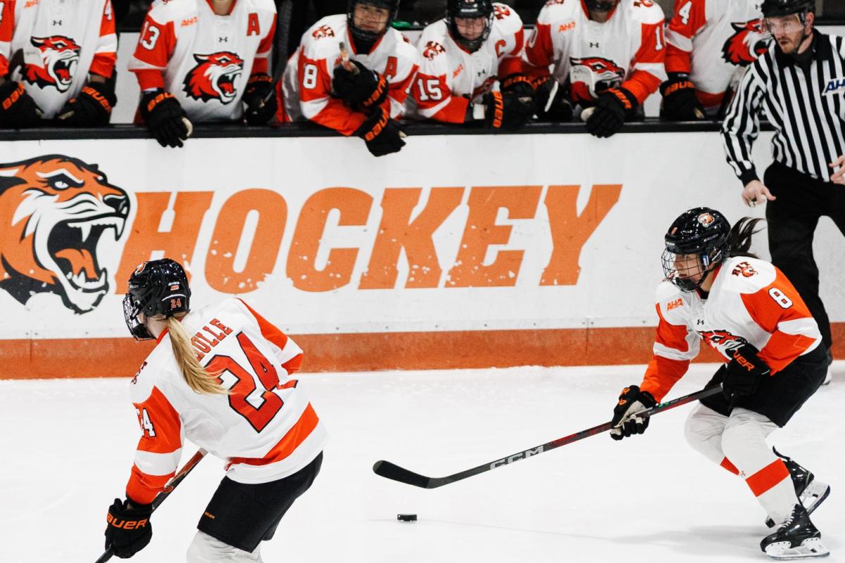 Two RIT women’s hockey players pursue the puck on the ice while teammates lean over the boards and watch the play from the bench area.