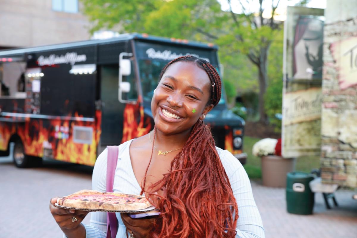 Participant holds a slice of pizza while standing near a food truck at an outdoor campus event with trees and event activities in the background.