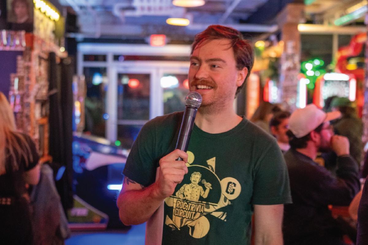Trivia host stands with a microphone in a busy entertainment venue, addressing participants seated throughout the space.