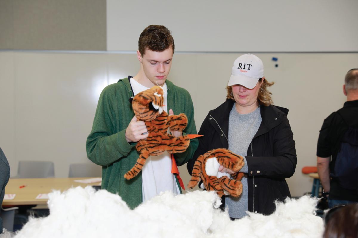 Two volunteers prepare tiger plush mascots, surrounded by piles of stuffing in a large event space.
