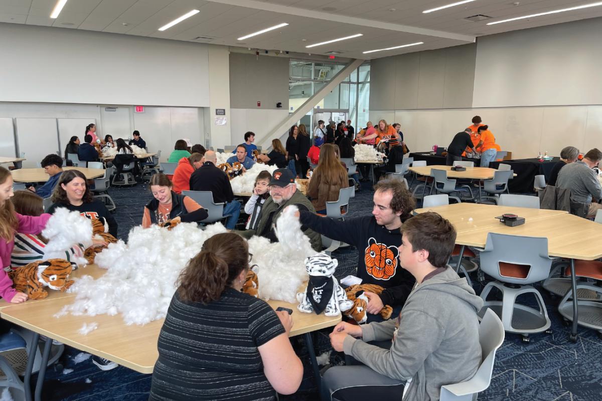 Volunteers and community members gather around tables stuffing plush tiger mascots in a large meeting room.