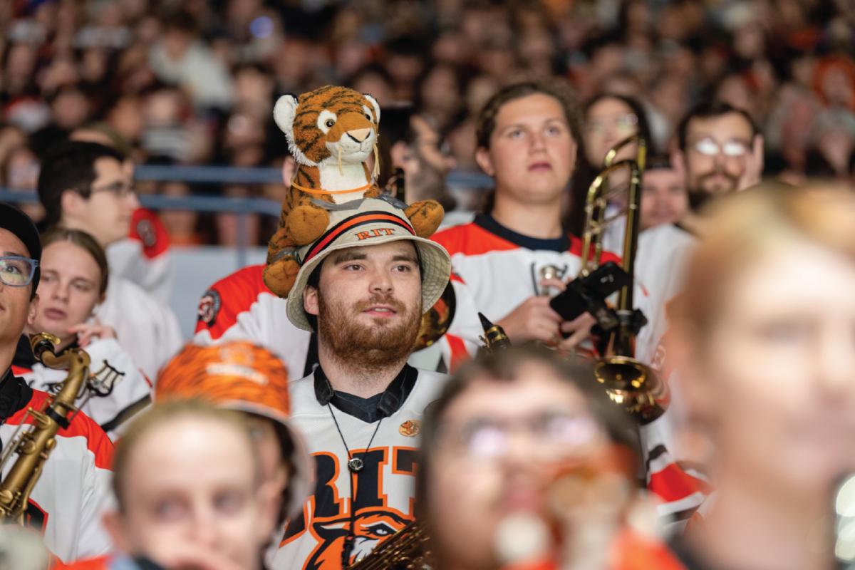 RIT Pep Band member wearing a tiger hat sits among fellow musicians in the arena stands during a hockey game.