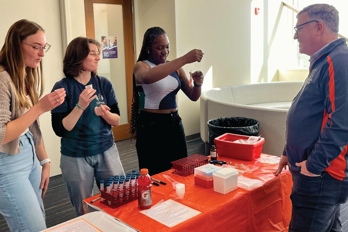 Four people gathered around a table with lab supplies, including test tubes and pipettes, as one person holds up a test tube during a hands‑on science activity.