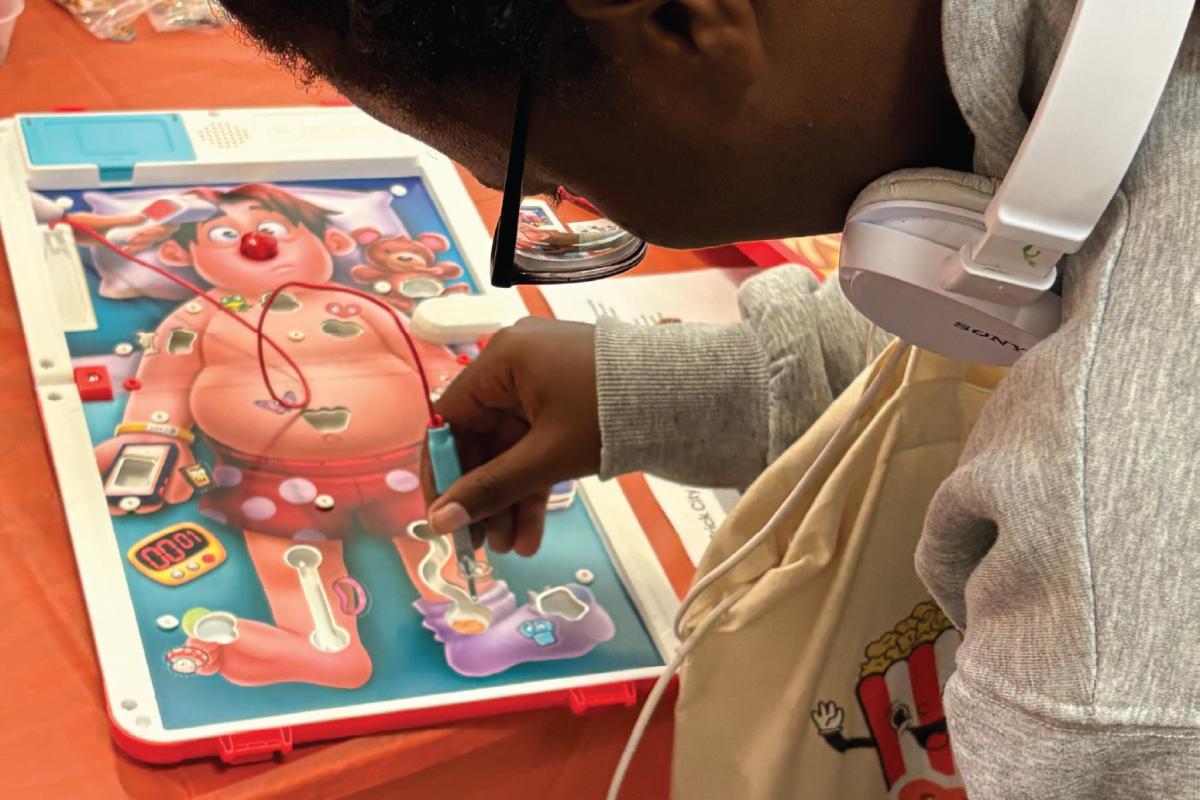 Person playing the Operation board game, using metal tweezers to remove a piece while wearing glasses and headphones.