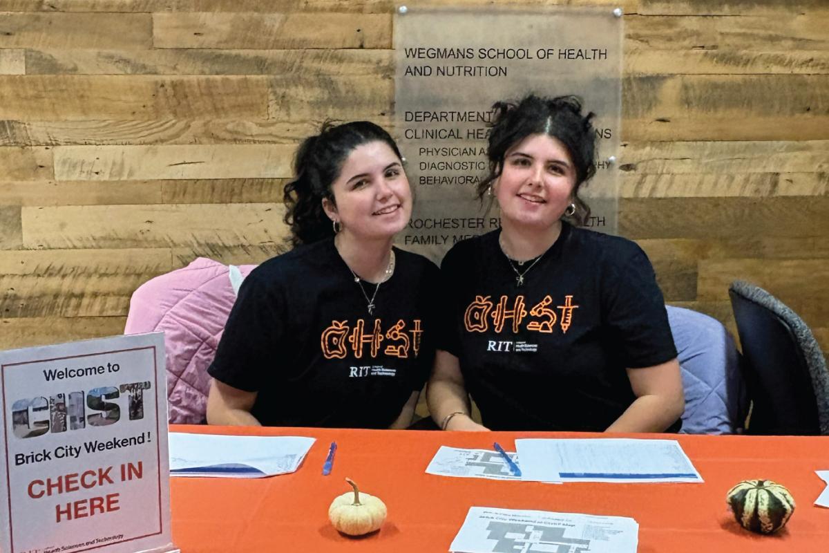 Two CHST students wearing the limited-edition College of Health Sciences and Technology t-shirts sit behind a Brick City Weekend check-in table with registration materials and a "CHST Brick City Weekend Check In Here" sign.