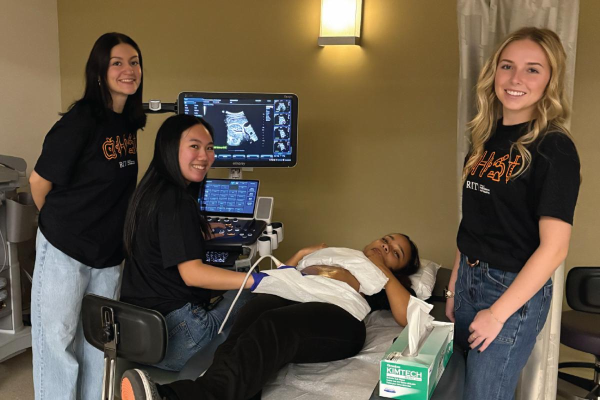 Four people in black RIT health sciences shirts observing an ultrasound exam as one person uses a transducer on a patient’s abdomen.