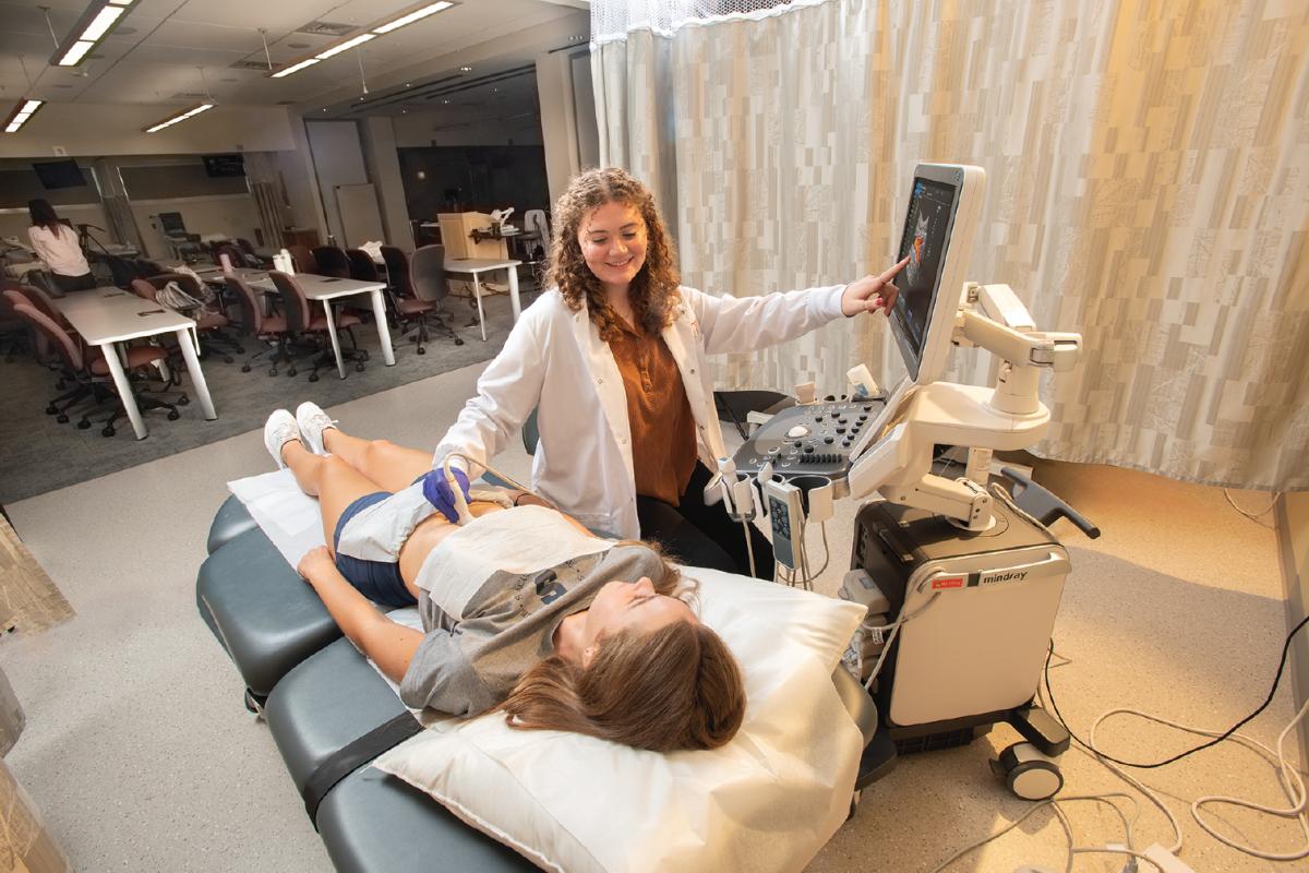 Person in a white lab coat performing an ultrasound on someone lying on an exam table while pointing to the image on the Mindray machine’s monitor.