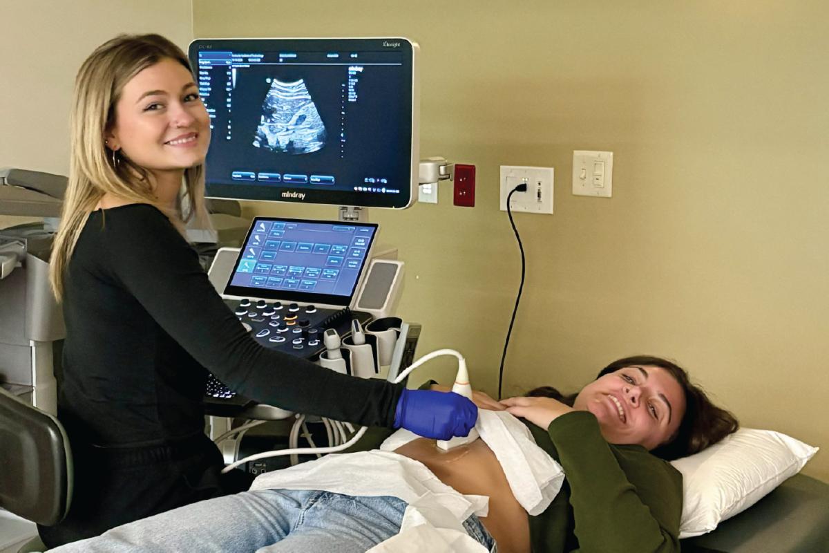 Person lying on an exam bed with a drape over the abdomen while another operates an ultrasound machine, the screen showing a grayscale scan.