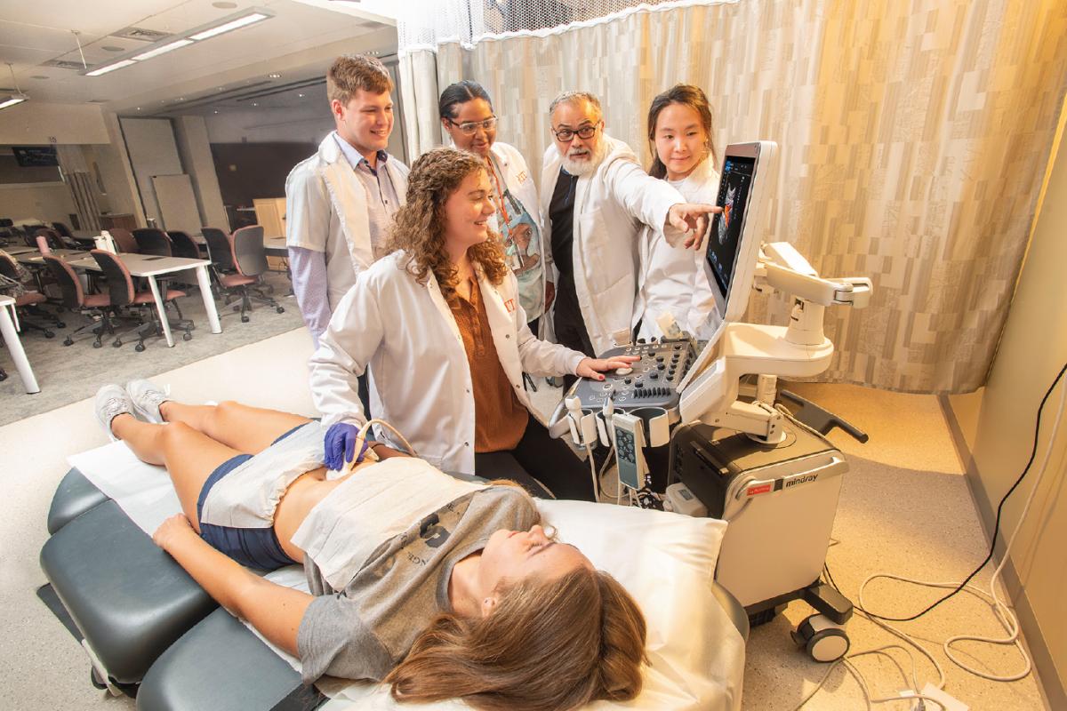 Group gathered around an ultrasound machine as a person in a lab coat scans someone lying on an exam table while another points at the monitor.