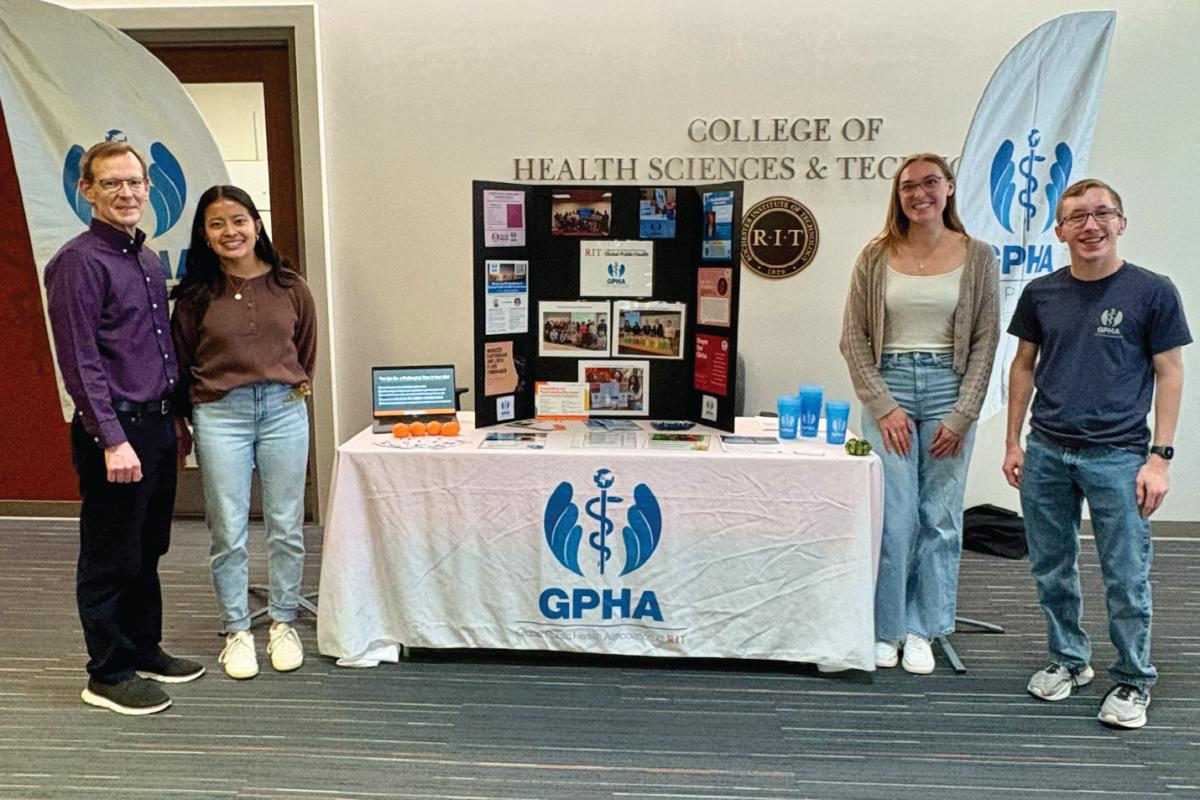 Four people standing beside a Global Public Health Association table with a display board, blue GPHA cups, and public‑health materials in an indoor RIT setting