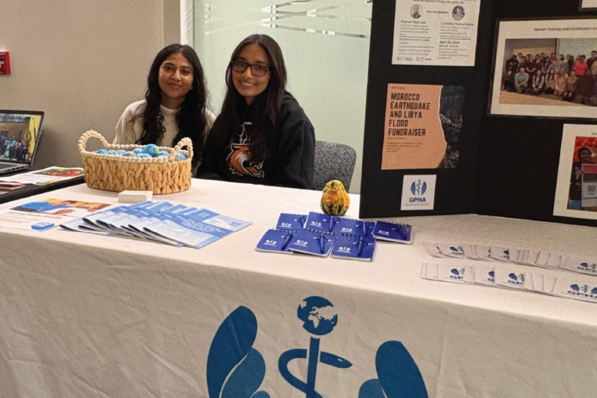 Two people seated behind a Global Public Health Association table with pamphlets, blue cards, and a display board featuring event posters and photos.