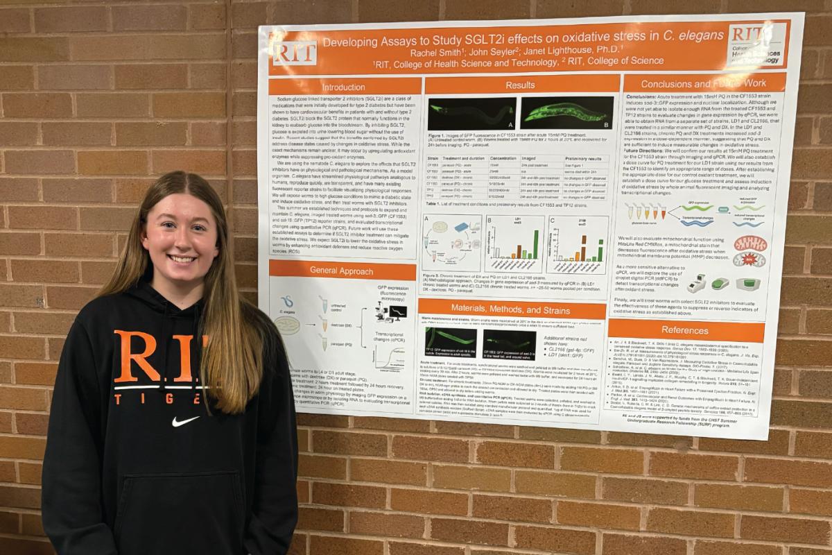 Person standing in front of a scientific research poster, wearing a black RIT Tigers hoodie, presenting findings with diagrams, graphs, and microscopy images.