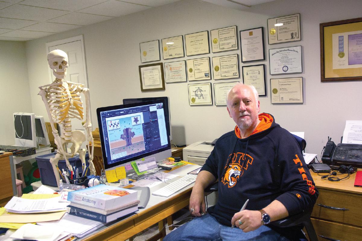 Person seated at a desk with a computer displaying a scientific illustration, surrounded by books and papers, with a full skeleton model standing nearby and framed certificates on the wall.