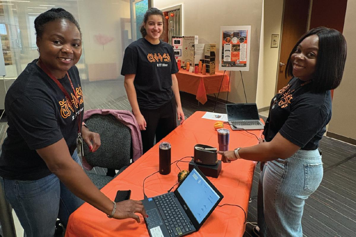 Three people in matching black RIT shirts standing around a table with laptops and a small electronic device, engaged in a Veggie meter demonstration at an indoor event