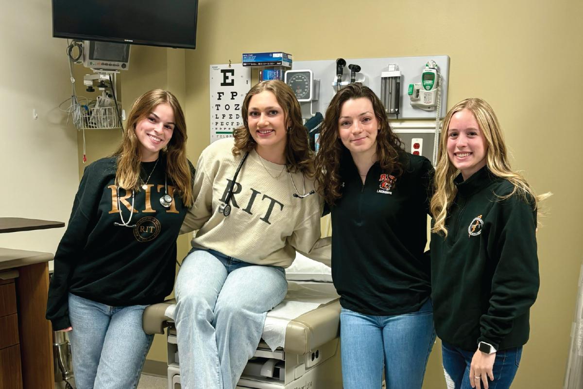 Four people in a medical exam room, one seated on the exam table and others standing nearby, with RIT clothing visible and typical clinical equipment on the walls.