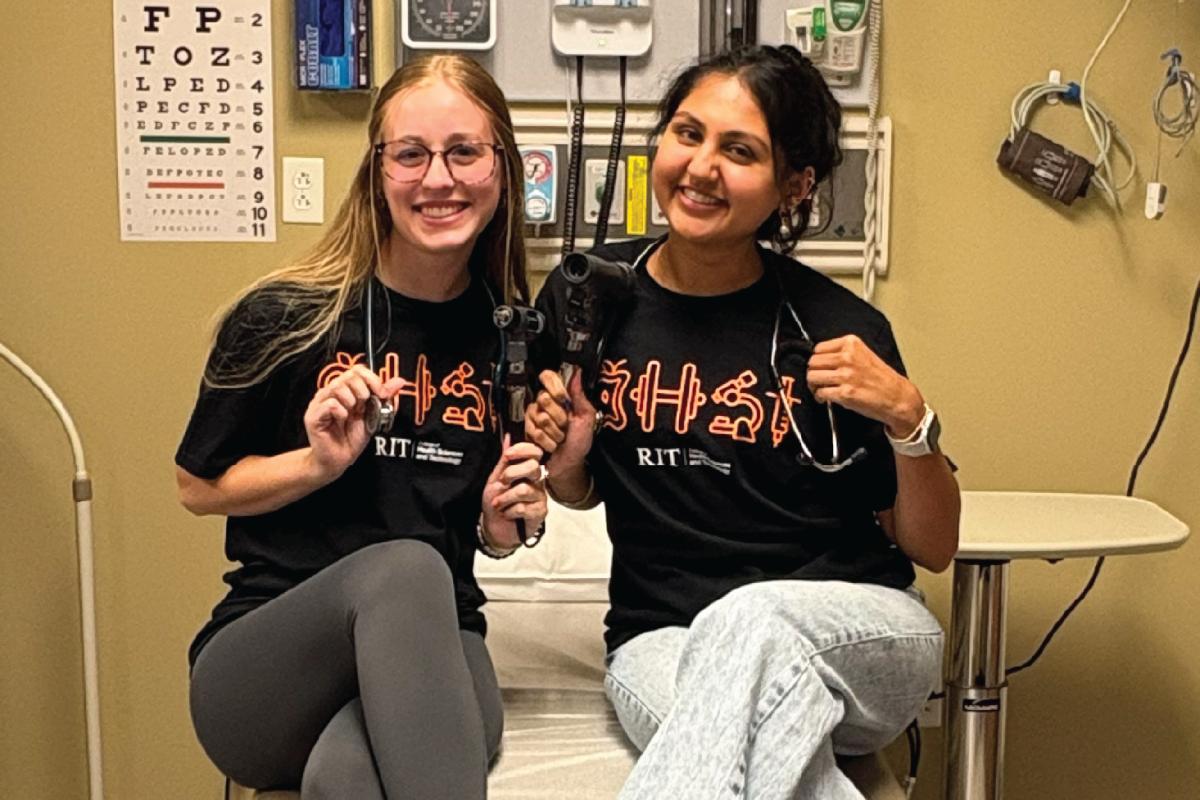 Two people sitting on an exam table wearing black RIT health sciences shirts, each holding an otoscope with stethoscopes around their necks, surrounded by wall‑mounted medical tools.