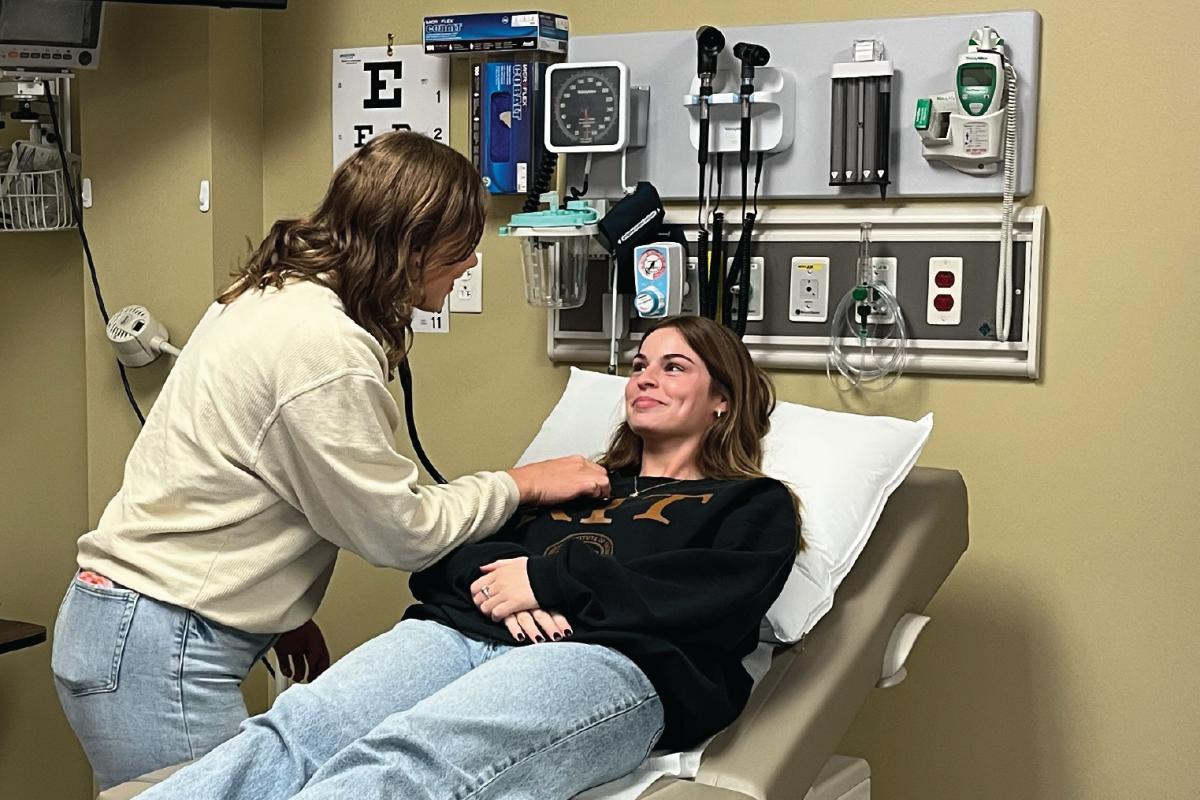 Person using a stethoscope to listen to someone lying on an exam table in a medical room with standard diagnostic tools and an eye chart on the wall.