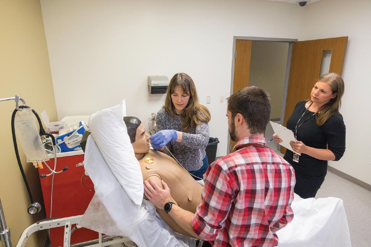 Person inserting a tube into a medical training mannequin’s nose while another observes and a third supervises with a clipboard in a clinical simulation room.