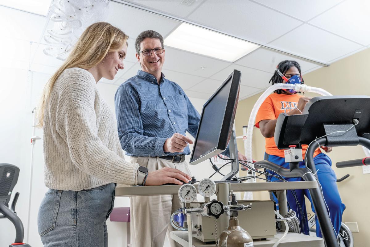 Person on a treadmill wearing a breathing mask while two researchers review data on a nearby computer in a bright exercise‑science lab.
