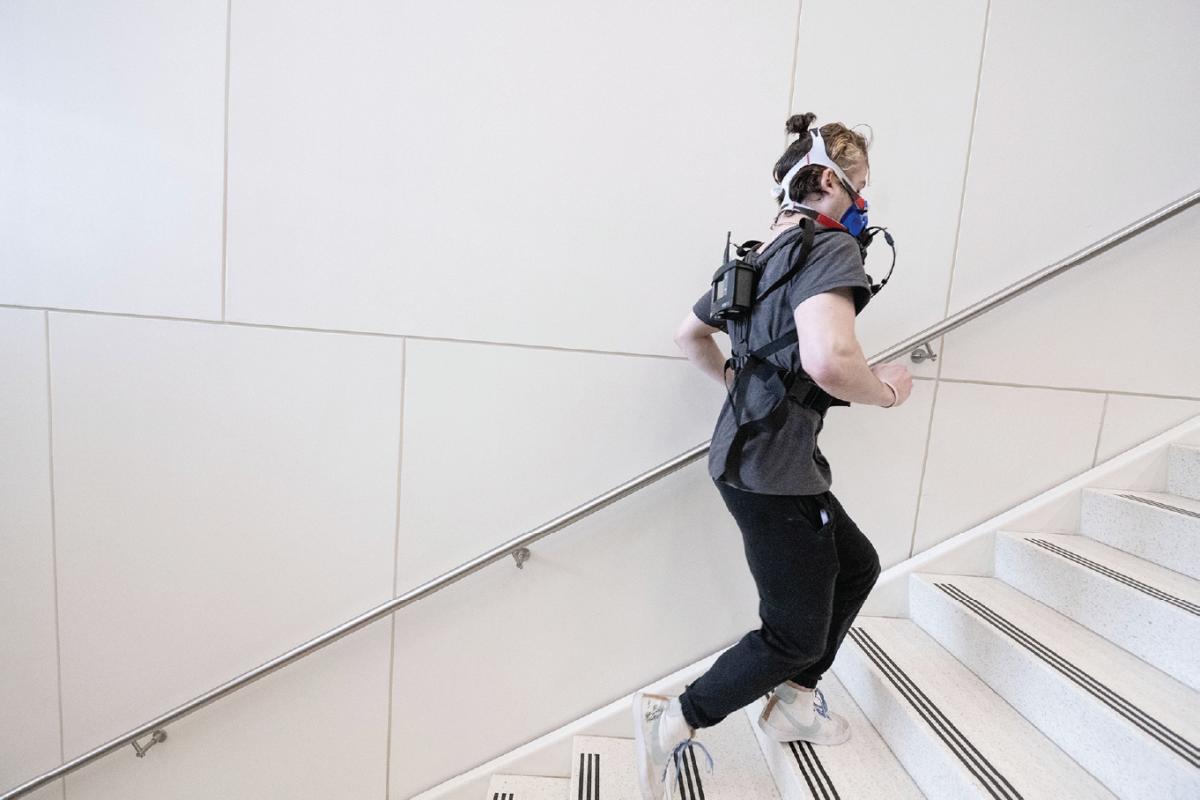 Person climbing indoor stairs wearing a harness, backpack‑mounted sensors, and a respiratory mask with tubing, participating in a monitored exercise or physiology test.