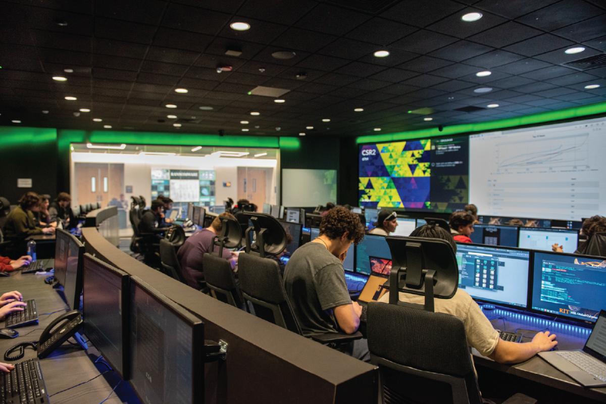 Participants work at computer stations in a cybersecurity lab during a hackathon, with coding screens, large digital displays, and collaborative workspaces throughout the room.